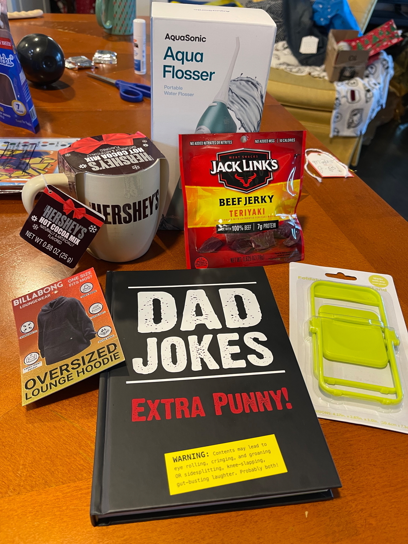 A variety of gift items including a mug, a hoodie package, a book titled “Dad Jokes,” a water flosser, beef jerky, and a mobile phone stand are arranged on a table..