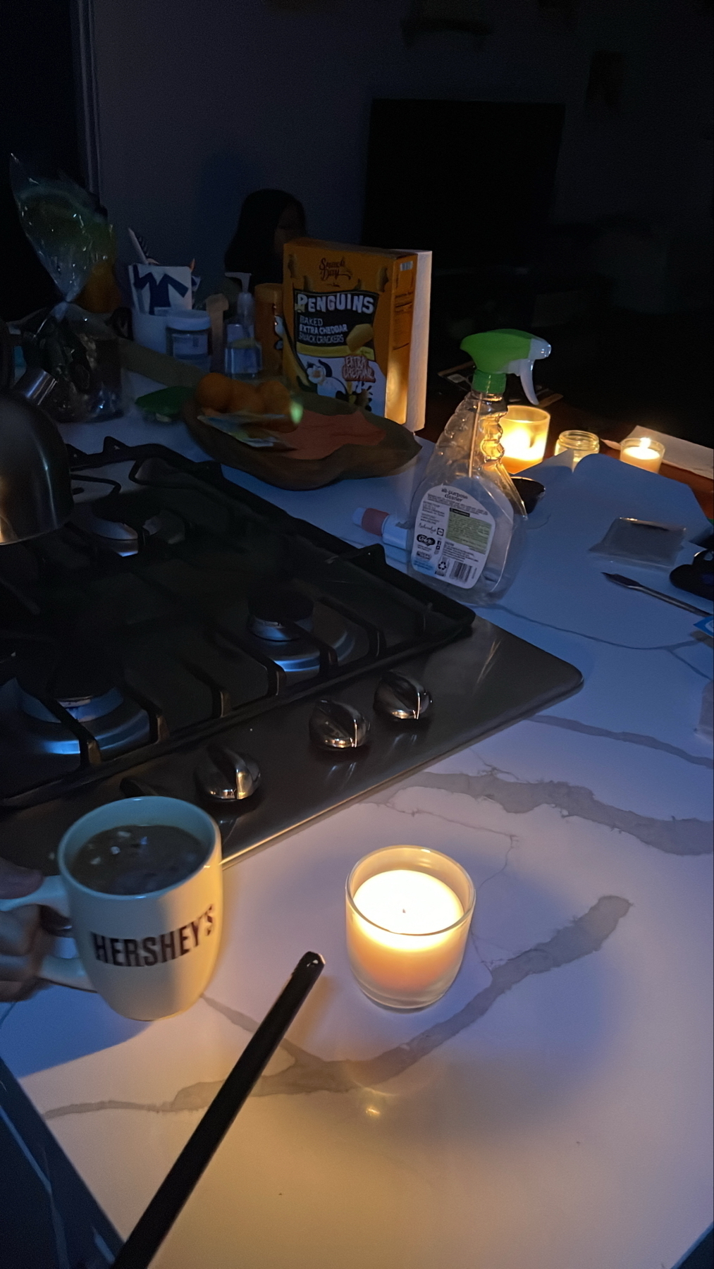 A dimly lit kitchen features a Hershey’s mug being held near a lit candle on a countertop, with snacks and other candles in the background.