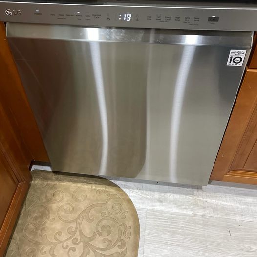A brand new LG dishwasher with a stainless steel finish is shown surrounded by wood cabinets and a LVM faux whitewashed wooden floor.