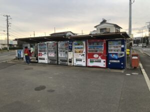 A row of beverage vending machines under a shelter in a parking lot in suburban Hiratsuka, Japan. A person and a child are seen buying drinks.